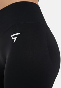 Črne visokokakovostne legice iz raztegljive tkanine z gladko teksturo. Imajo belo oznako, ki je izpostavljena ob strani.