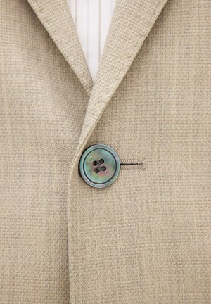 Beige blazer met textuur, vastgemaakt met een grote iriserende viergaatjesknop, gedragen over een wit shirt met verticale strepen.