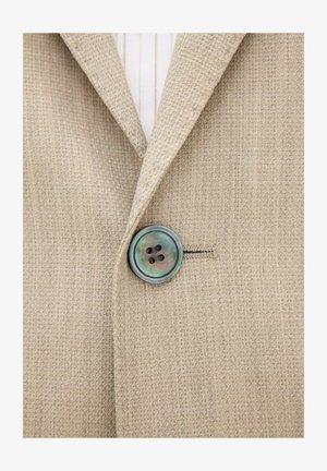 Blazer beige strutturato con un unico grande bottone iridescente a quattro fori, abbottonato sopra una camicia bianca a righe verticali.