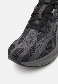 Chaussure de sport noire et grise avec un dessus en tricot texturé orné de rayures diagonales, un laçage et une semelle en caoutchouc avec des stries.