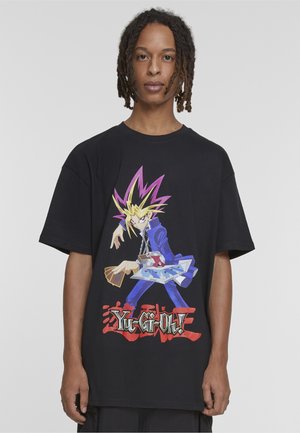Person trägt ein schwarzes oversized T-Shirt mit einem bunten Yu-Gi-Oh!-Charakter, der Karten und einen Duell-Disck hält, mit rotem Logo-Text darunter.