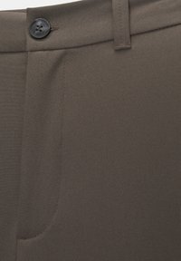 Gros plan sur un pantalon marron montrant la ceinture, un bouton noir, une passants de ceinture et une couture partielle de poche avant.