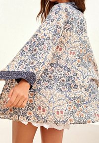 Luźna kimono w kwiatowy wzór z kremowego materiału w niebieskie, różowe i zielone akcenty; szerokie rękawy oraz koronkowe wykończenie u dołu.