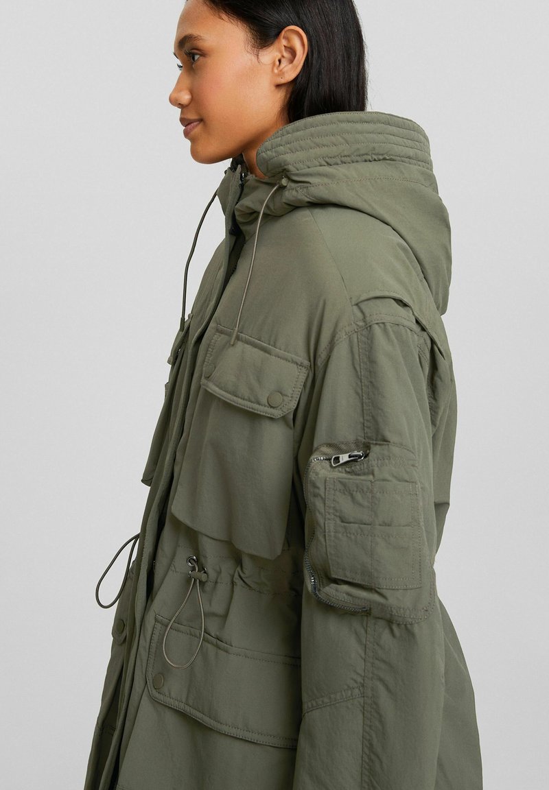 Parka olive avec capuche, poches avant, poche à glissière sur la manche et cordon de serrage à la taille, fabriquée dans un tissu lisse et durable.
