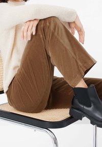 Bruna kordbyxor med en texturerad yta, kombinerat med svarta ankelstövlar med elastiska sidor, sittande på en vävd stol.