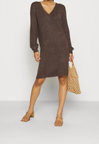 Robe en tricot marron avec un col en V et des manches longues, présentant une coupe décontractée. Associée à un sac tissé beige et des sandales à talons carrés couleur fauve.