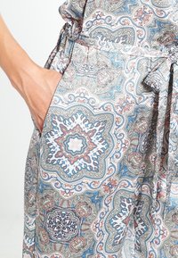 Main dans la poche d'un vêtement en coton avec un motif floral et géométrique complexe en bleu, rouge et beige, et une ceinture en tissu nouée.