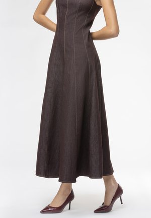 Abito maxi in denim marrone con cuciture verticali e una parte superiore aderente, abbinato a scarpe con tacco alto bordeaux. Tessuto liscio, silhouette ad A.