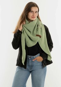 Groene gebreide sjaal met een textuurpatroon, om de nek van een model gedrapeerd. Bevat decoratieve hardware en een bruine label. Draagt met een zwarte jas.