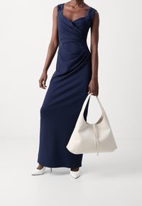 Marineblauwe, vloerlength jurk met kantaccenten op de schouders, gedrapeerd ontwerp, gecombineerd met een witte leren tas en witte puntige hakken.