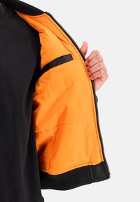 Osoba držící otevřenou černou bundu, která odhaluje jasně oranžové prošívané podšívky s černou kapsou na západku uvnitř.