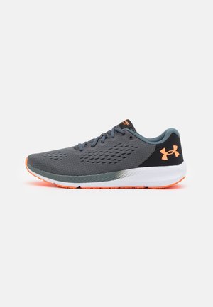 Zapatilla de correr gris oscuro y negra con el logo naranja de Under Armour, acentos naranjas en la suela y entresuela acolchada blanca, mostrada sobre fondo blanco.