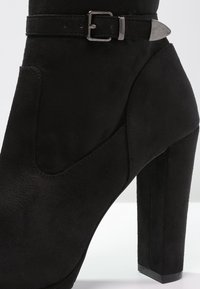 Stivaletto nero in suede con tacco a blocco alto. Presenta una fascia decorativa con fibbia e una texture liscia, evidenziando una silhouette moderna.