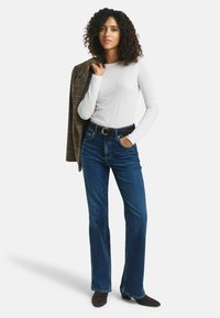 Blauwe spijkerbroek met flared pijpen en een hoge taille, gecombineerd met een wit top met lange mouwen. Een geruit blazer hangt over de schouder. Bruine enkelhoge laarzen.