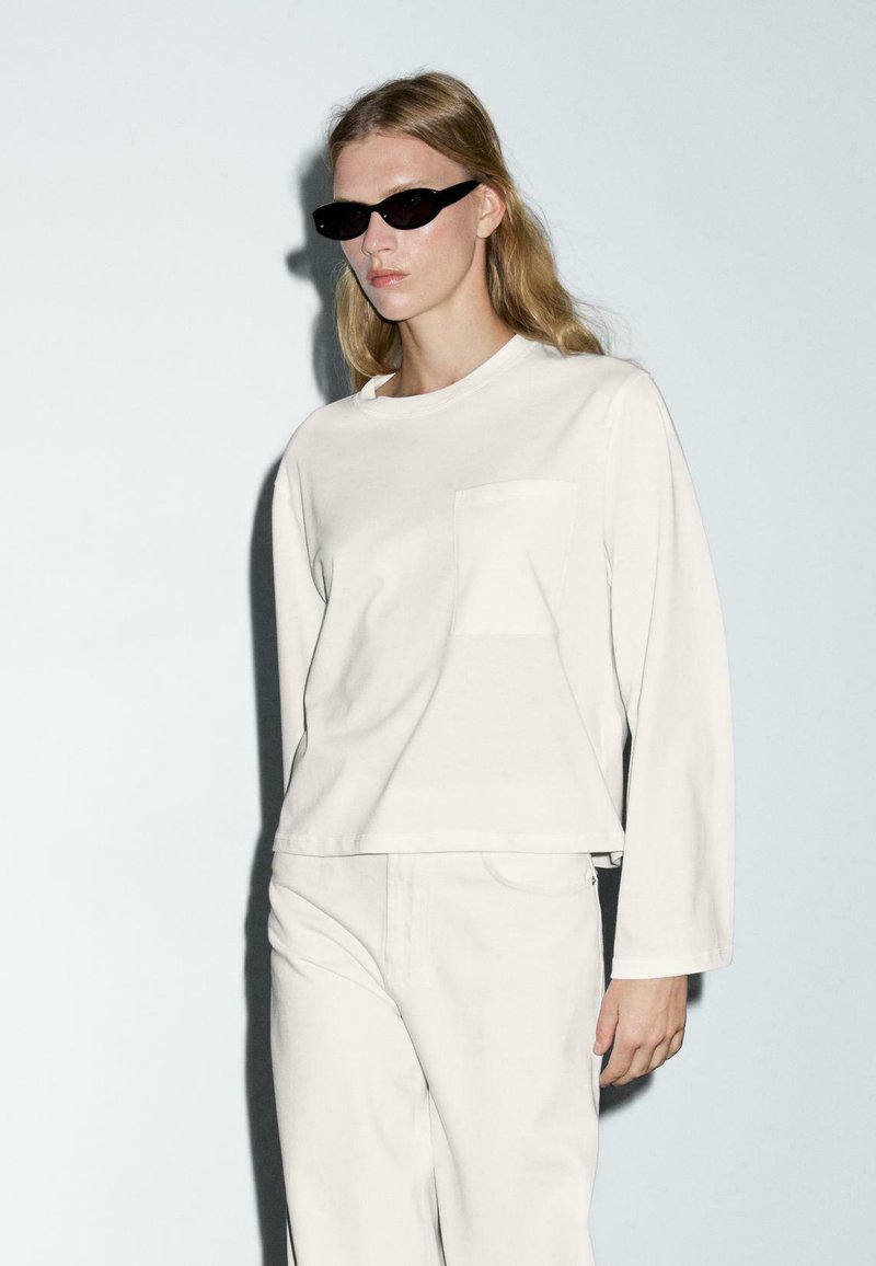Witte longsleeve t-shirt met een voorzak, gestyled met bijpassende lichtgekleurde broek. Het model draagt zwarte zonnebril tegen een lichte achtergrond.