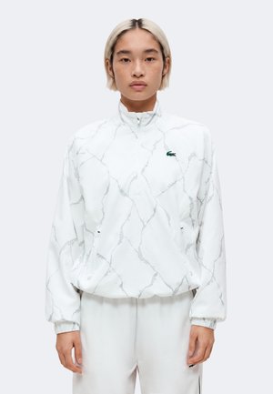 Persoon met kort blond haar die een witte zip-up jas met marmerpatroon en groen krokodillenlogo draagt, en bijpassende witte broek.
