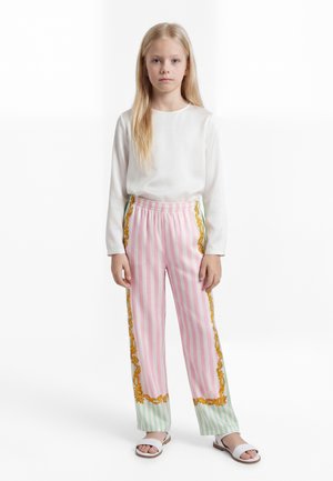 Jauna mergaitė su ilgais šviesiais plaukais, vilkinti baltą ilgomis rankovėmis palaidinę, rausvai žalias dryžuotas kelnes ir baltus sandalai, stovinti ramiai.