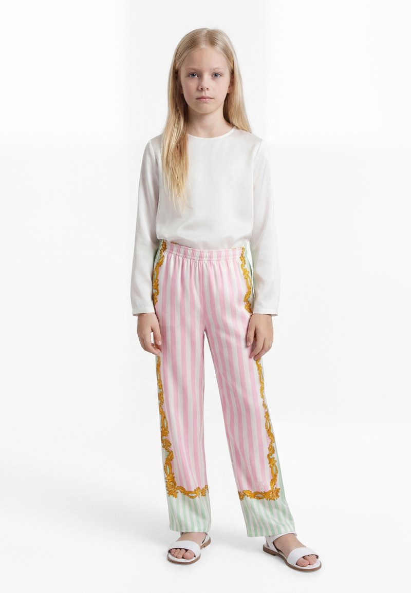 Mladé dievča s dlhými blond vlasmi, oblečené v biele tričko s dlhým rukávom, ružovo-zelené pruhované nohavice a biele sandále, stojí nehybne.