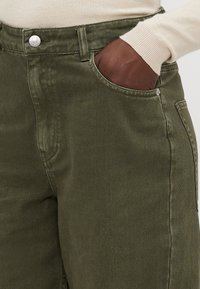 Olive krāsas denim bikses ar augstu jostu, sānu kabatām un sudraba pogu akcents. Audumam ir gluda tekstūra un viegla spīduma.