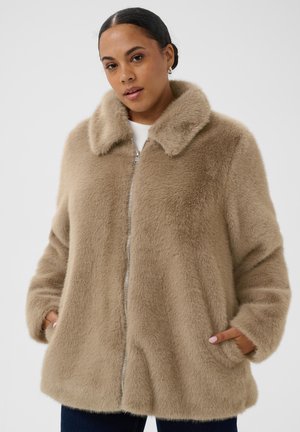 Femme portant un manteau zippé beige duveteux, les mains dans les poches, sur un fond clair uni.