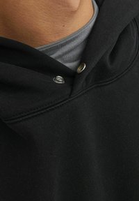 Sort hættetrøje med en glat tekstur, prydet med sølvknapper og en lagdelt grå T-shirt under.