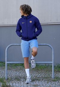 Marine blauwe zip-up hoodie met een logo, blauwe denim shorts, witte sokken en grijze sneakers, geposeerd tegen een reling in een buitenomgeving.