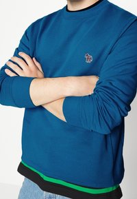 Blauwe trui met geribde manchetten en halslijn, voorzien van een klein geborduurd paardenlogo. Gelaagde zoom met zwarte en groene details.