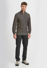 Brauner Strickpullover mit Reißverschluss am Hals, kombiniert mit schwarzen Hosen und grauen Schuhen. Hat lange Ärmel und ein strukturiertes Muster.