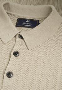 Beige kötött pólóing, texturált hullámos mintával, gallérral, három fekete gombbal és belső varrással ellátott márkacímkével.