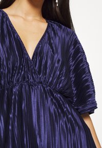 Lila plisserad klänning med djup V-ringning och korta, breda ärmar. Tyget har en blank, strukturerad yta och samlas vid midjan.