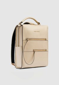 Zaino beige con design strutturato, dettagli in metallo color oro, multiple tasche con cerniera e manico superiore. Materiale liscio con accenti neri a contrasto.