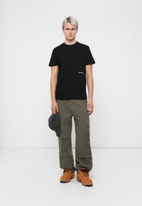 Zwarte katoenen t-shirt met een klein wit logo, gecombineerd met olijfgroene cargobroeken en tanlaarzen. Het model houdt een grijze pet vast. Neutrale achtergrond.