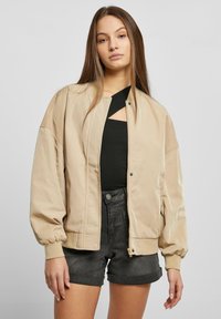 Giacca bomber beige con una texture liscia, polsini a coste e zip frontale. Indossata sopra una maglietta nera, abbinata a pantaloncini neri.
