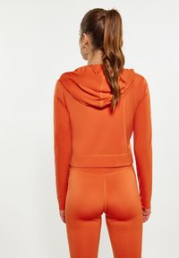 Giacca arancione con cappuccio, a maniche lunghe, realizzata in materiale liscio; abbinata a leggings attillati coordinati. Visibile il dettaglio delle cuciture.