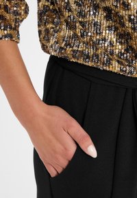 Blouse à paillettes dorées et noires à manches courtes, associée à un pantalon noir avec une poche latérale et des plis. Texture lisse et finition brillante.