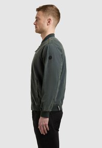 Homme en veste bomber verte et pantalon noir, debout de profil face à gauche sur un fond uni.