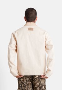 Lichtbeige jack van gestructureerde stof, met een kraag en een rechthoekig label op de achterkant. Gecombineerd met patronen cargobroek.