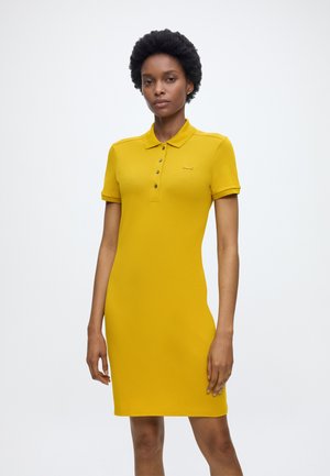 Femme aux cheveux courts et bouclés, vêtue d'une robe polo jaune moutarde à manches courtes, debout devant un fond clair uni.