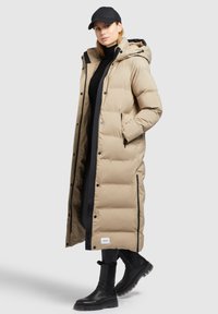 Beige lang pufferjakke med hætte, sorte accenter og lynlås. Modellen har en sort turtleneck og kasket på, parret med sorte knæhøje støvler.