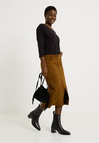 Top noir à manches longues, jupe midi en velours côtelé marron avec fente sur le côté, bottines noires et petit sac en suède noir avec des franges.