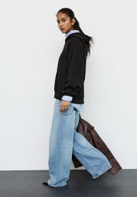 Černá mikina s oversized střihem navrstvená přes světle modrou košili s knoflíky, doplněná širokými světle modrými džínami. Hnědá kožená bunda přehozená zezadu.