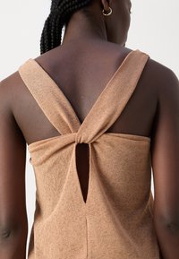 Femme aux cheveux tressés portant un haut beige sans manches avec des bretelles croisées et une petite découpe en forme de trou de serrure dans le haut du dos.