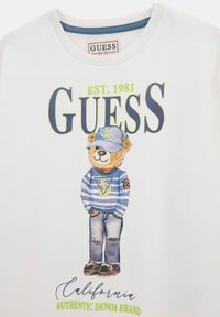 T-shirt bianco con una grafica di un orso in un maglione a righe blu e cappello, abbinato a jeans in denim e scarpe marroni. Logo "GUESS" incluso.