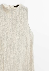 Haut blanc sans manches à texture, avec un col rond haut et une fermeture par boutons au dos, étalé à plat sur une surface blanche.