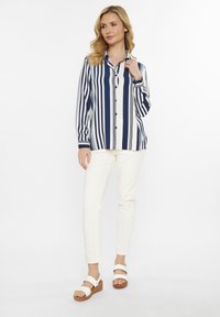 Camicia a righe navy e bianche con colletto, abbinata a pantaloni bianchi slim-fit e sandali bianchi su una piattaforma marrone testurizzata.