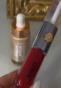 Röd läppstift i en klar tub med ett silverlock, med en guldlogotyp. En glasflaska med upplysande serum och en vit pipett finns i bakgrunden.