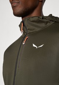 Ελαιόχρωμο μπουφάν με ψηλό γιακά, που διαθέτει πορτοκαλί  κορδόνια, λευκό λογότυπο και λεία υφή. Ελαφρύ, ιδανικό για χρήση σε εξωτερικούς χώρους.