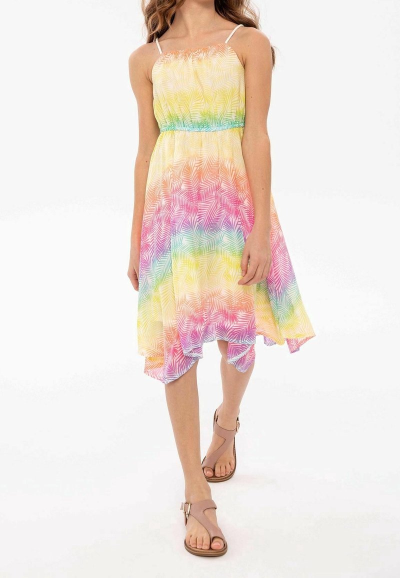 Fille portant une robe arc-en-ciel multicolore sans manches avec des motifs de feuilles et des sandales beige à bouts ouverts, debout sur un fond blanc.