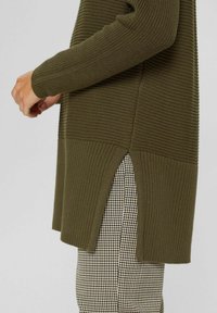 Vue latérale d'une personne portant un pull en maille côtelée vert olive avec une fente latérale, sur un pantalon à carreaux noir et blanc.