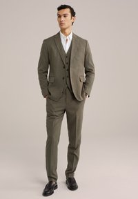 Öltöny világos olíva zöld szövetből, textúrázott mintával. Fitted zakó, mellény és slim lábujjú nadrág jellemzi. Fekete loaferek.